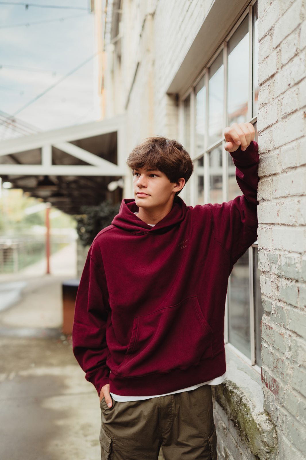 Senior boy in burgundy hoodie leaning against white brick wall at Revolution Mill in Greensboro with senior photographer Kelly McPhail Photography