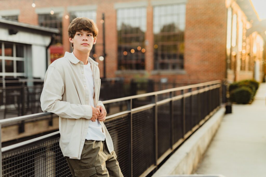 Senior boy in cream button-down shirt leaning on black metal railing at Revolution Mill with warm evening light, Greensboro senior photographer Kelly McPhail Photography