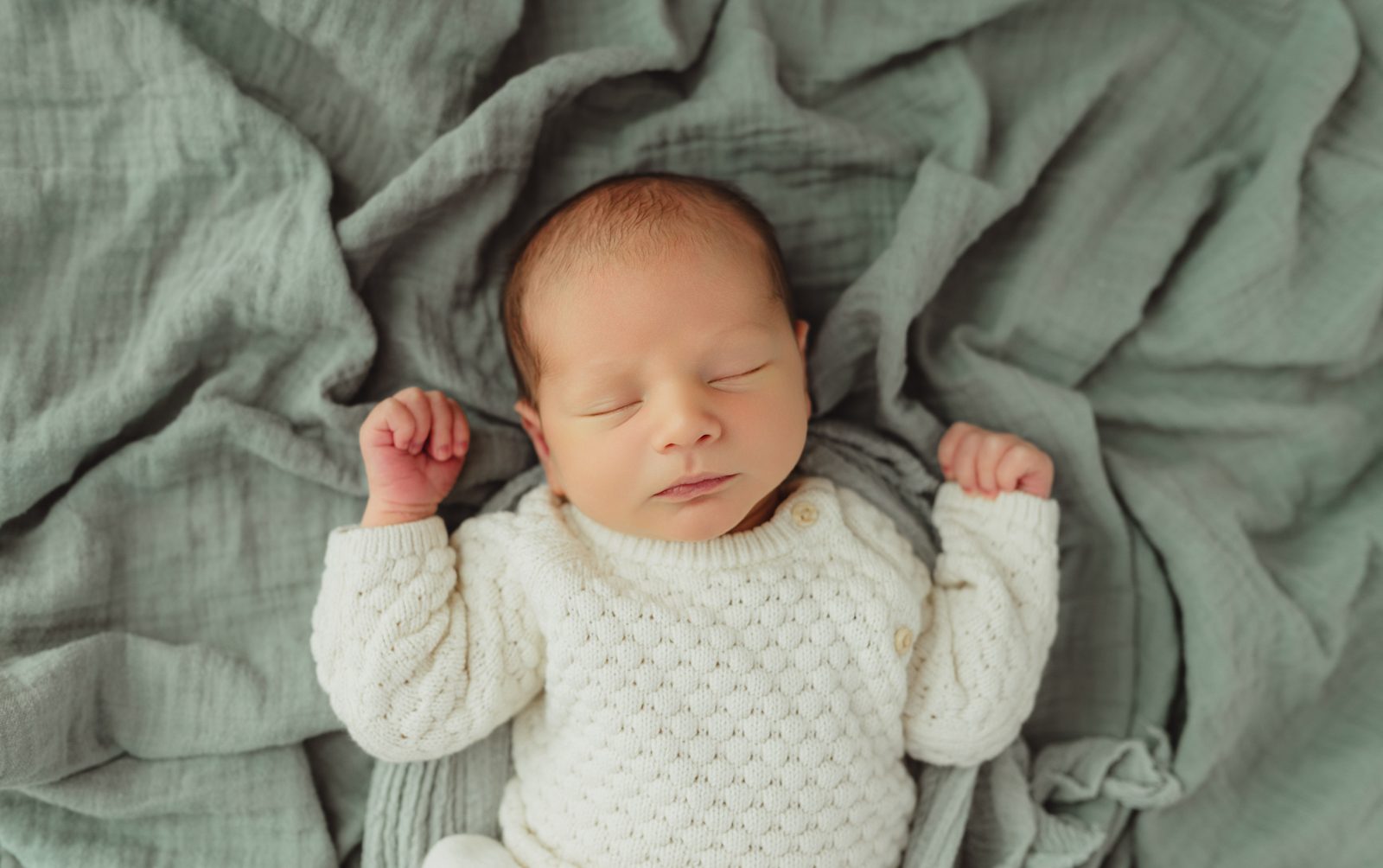 Sleeping newborn baby boy in cream knit sweater with fists raised on sage green muslin blanket — Greensboro newborn photography detail shot