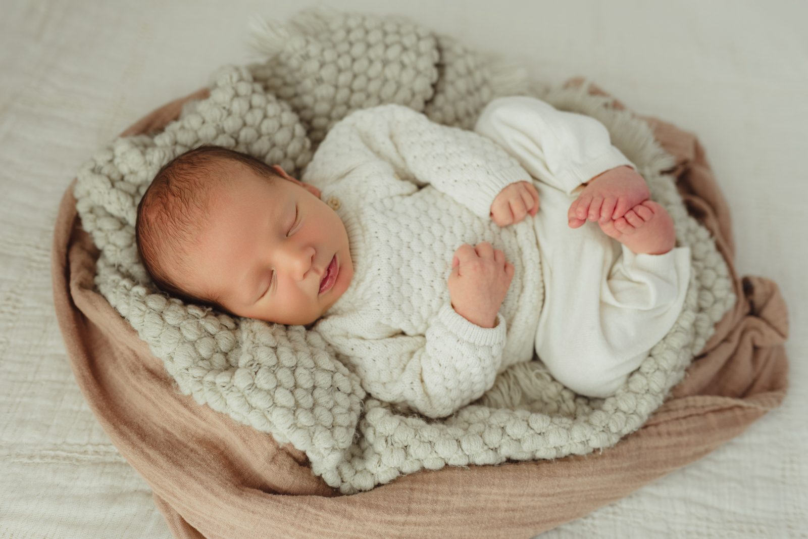 Newborn baby boy in cream knit tucked into nest of peach and cream blankets — tiny toes visible — Greensboro studio newborn session detail
