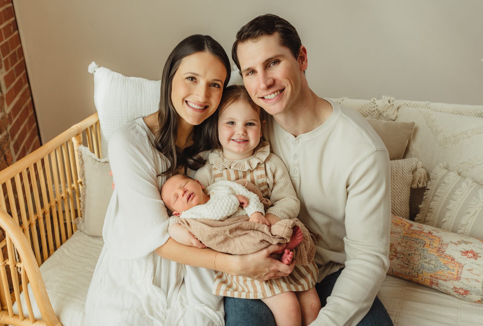 Family of four — mom, dad, big sister, and newborn baby boy — smiling together on rattan daybed at Revolution Mill Greensboro newborn studio