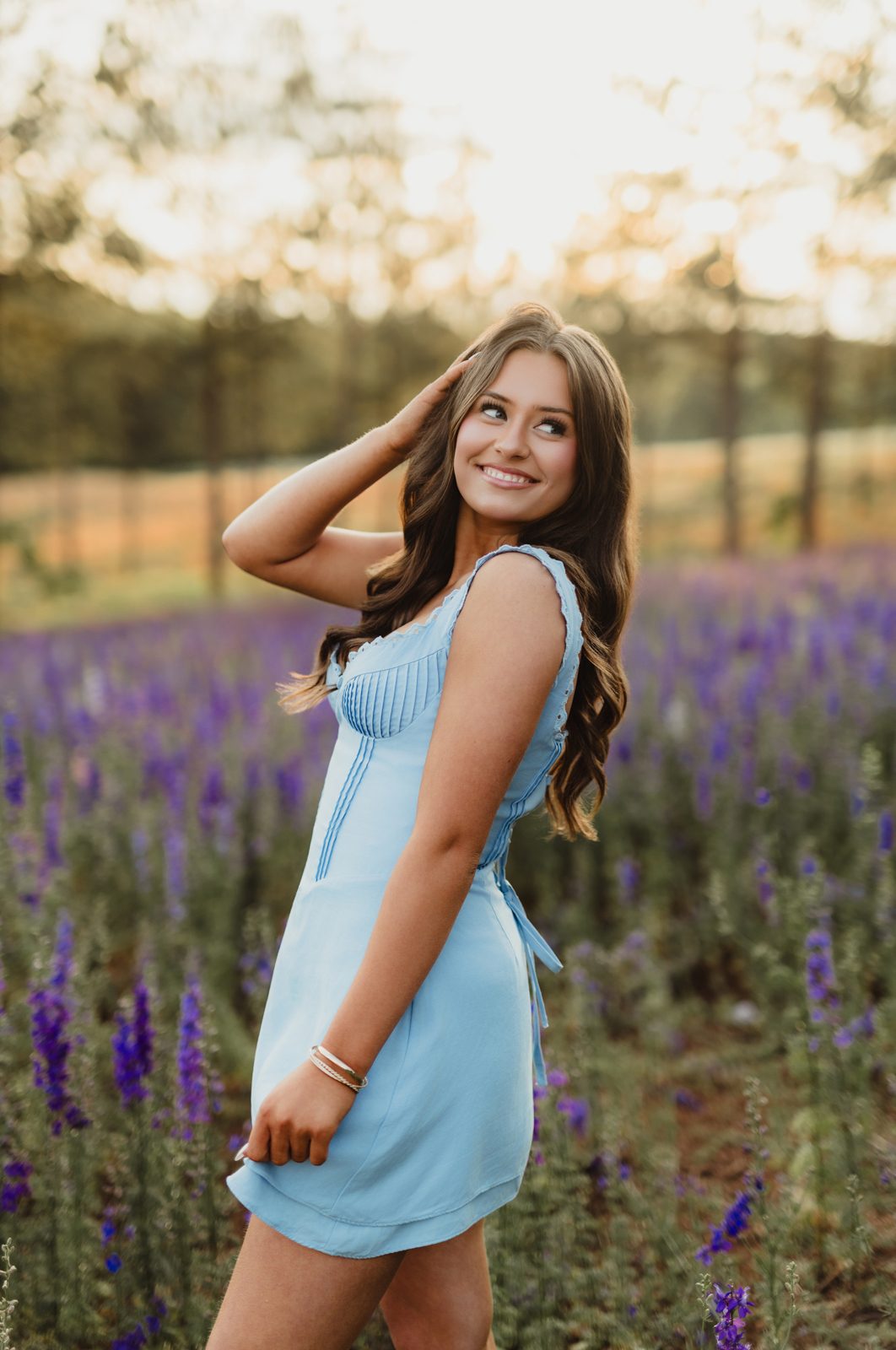 Senior in light blue pintuck mini dress standing in rows of purple larkspur at golden hour — Dogwood Farms Belews Creek NC senior photographer