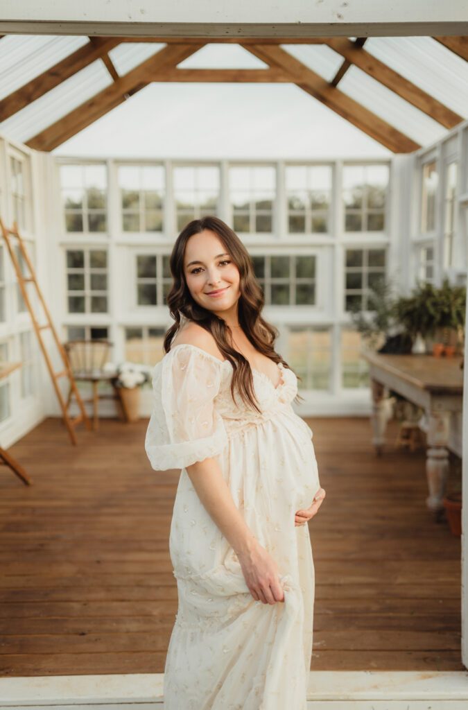Maternity Photos at The Lone Farm — A Greenhouse Session in Tobaccoville, NC-maternity photos Pregnant woman glancing over shoulder in cream dress inside The Lone Farm greenhouse Tobaccoville NC for maternity photos