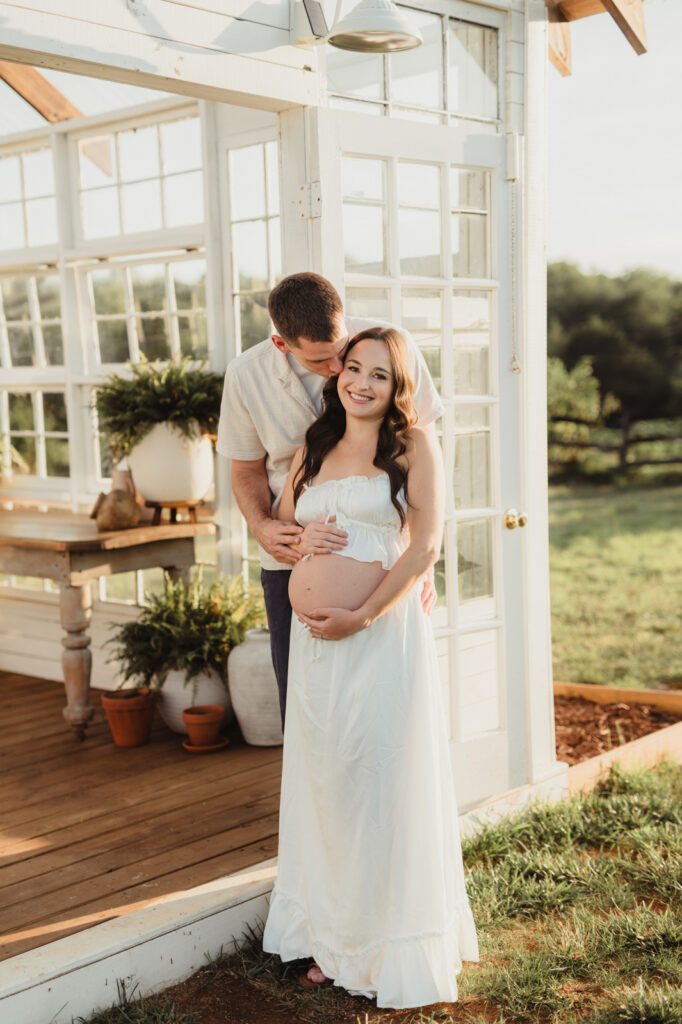 Maternity Photos at The Lone Farm — A Greenhouse Session in Tobaccoville, NC-maternity photos Husband kissing pregnant wife's forehead at greenhouse door during Lone Farm maternity photos near Greensboro