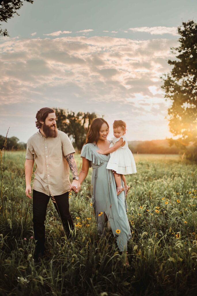 Parents holding hands with their toddler and baby daughter while walking through a grassy field during a Triad fall family photo session