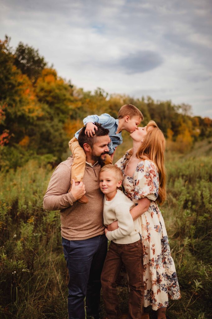 Father holding two sons while mother kisses their son in front of colorful peak fall trees during a Greensboro fall family photo session