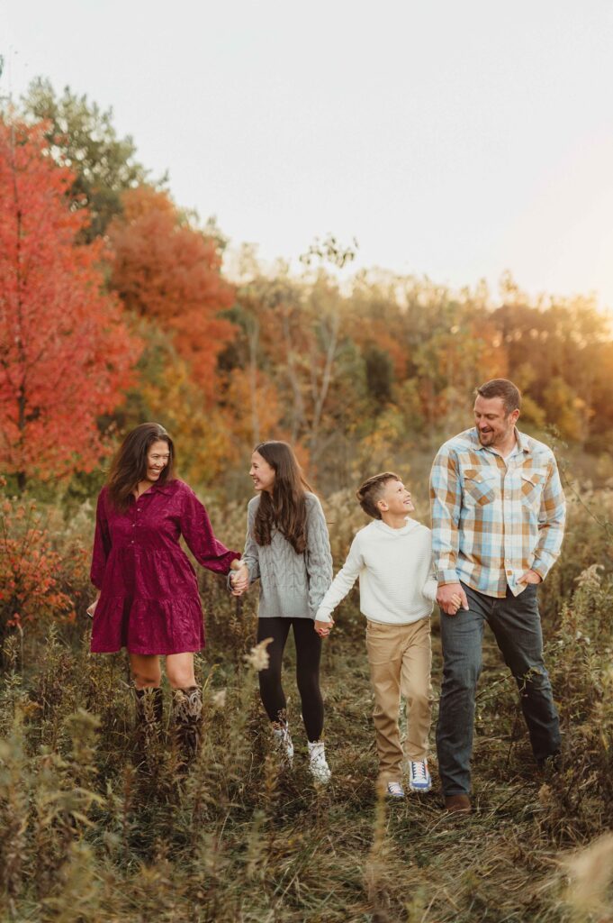 Family of four holding hands and walking through a field of red and orange fall foliage during a Greensboro fall family photo session