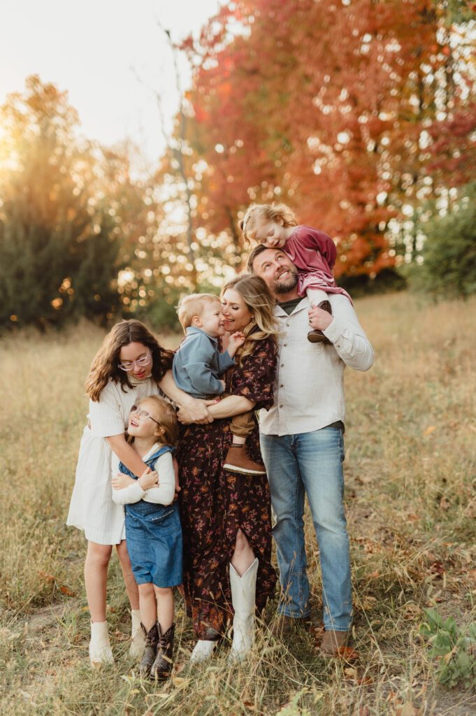 Family of six laughing and embracing together in front of red fall trees during a Triad fall family photo session