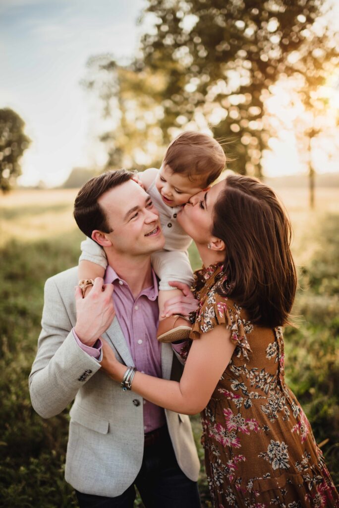 Toddler son on dad's shoulders while mom kisses her son on the cheek at sunset during Greensboro family photography session. 