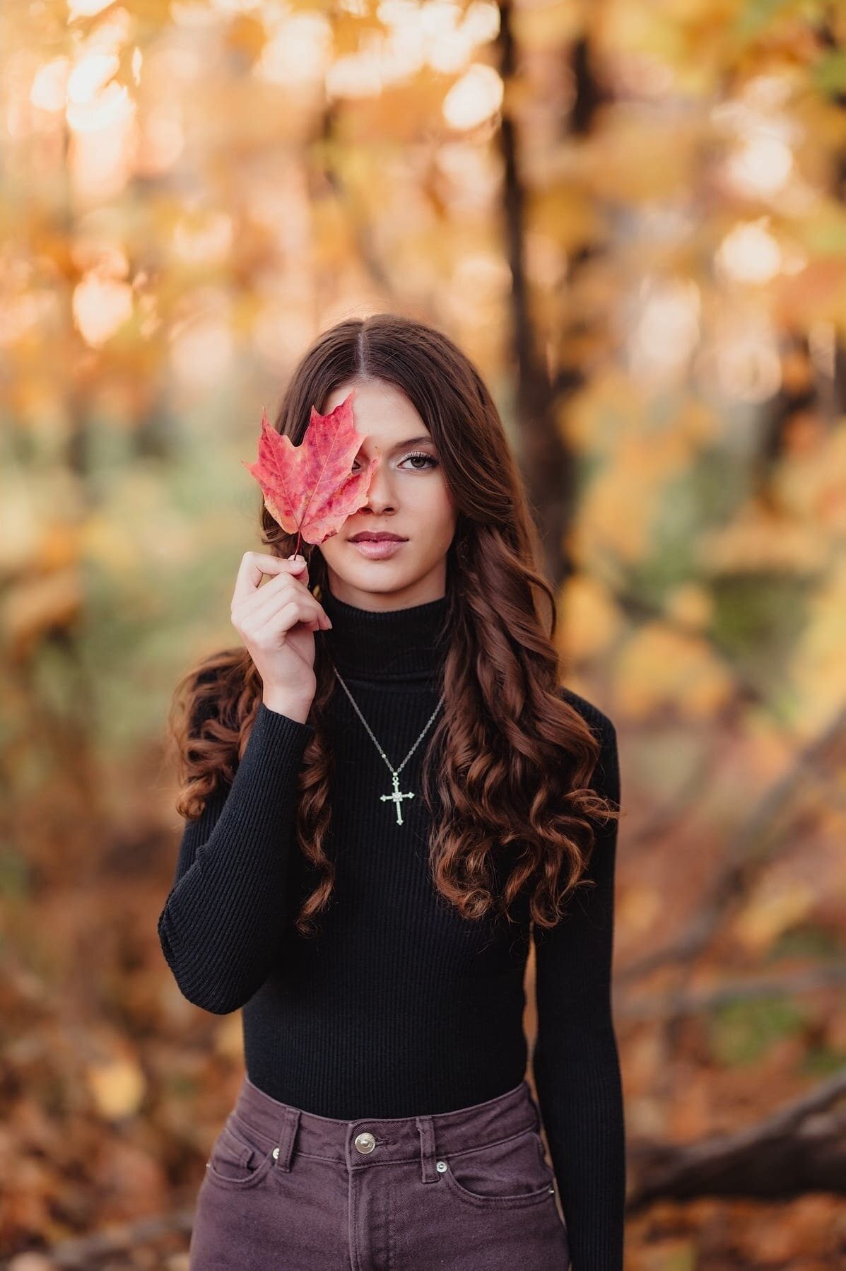 Senior girl holding a red maple leaf over one eye in an autumn forest, a nature-inspired senior session outfit idea near the Triad