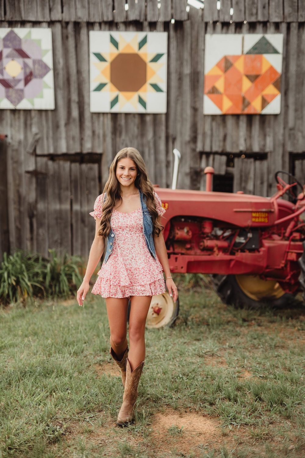 Senior girl in a floral dress, denim vest, and cowboy boots standing near a vintage tractor, a western senior session outfit idea