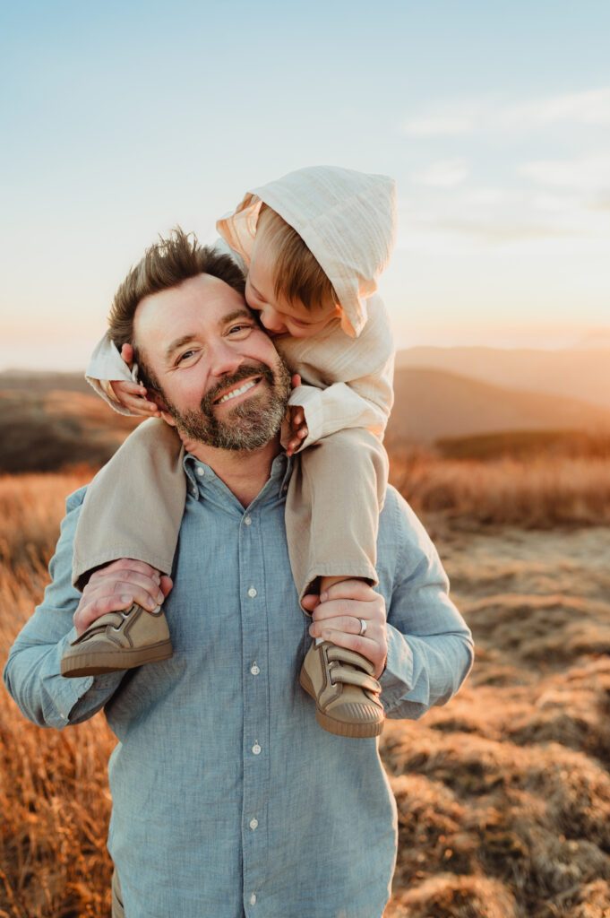 Father smiling while his toddler rides on his shoulders in a golden field during an NC fall family photo session