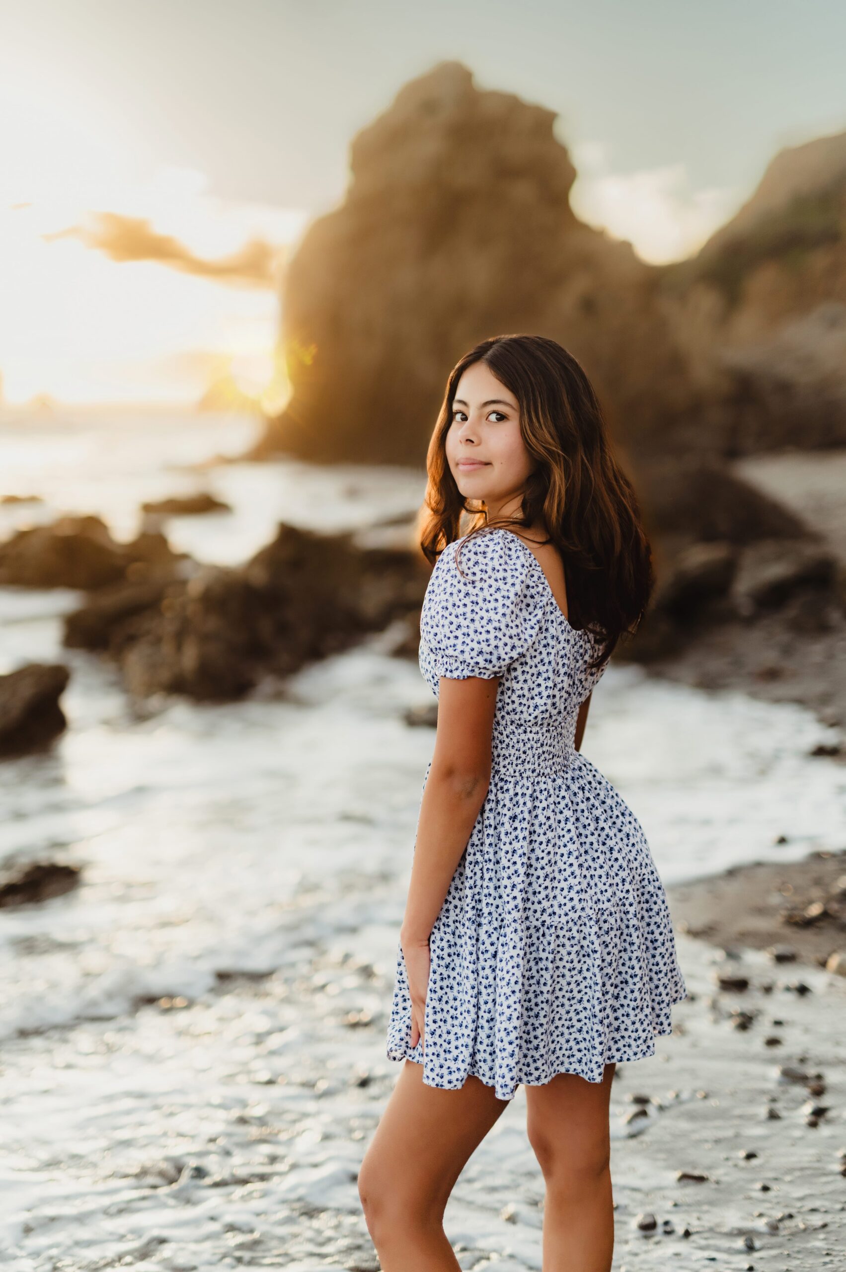 Senior girl in a linen floral dress looking back over her shoulder on the beach at sunset, a beachy senior session outfit idea