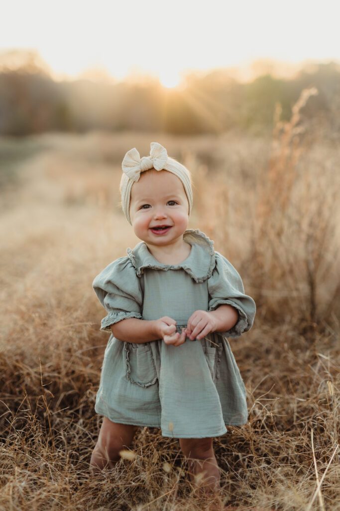 Toddler girl in a sage green dress and bow headband smiling in a golden field during a Triad fall family photography session