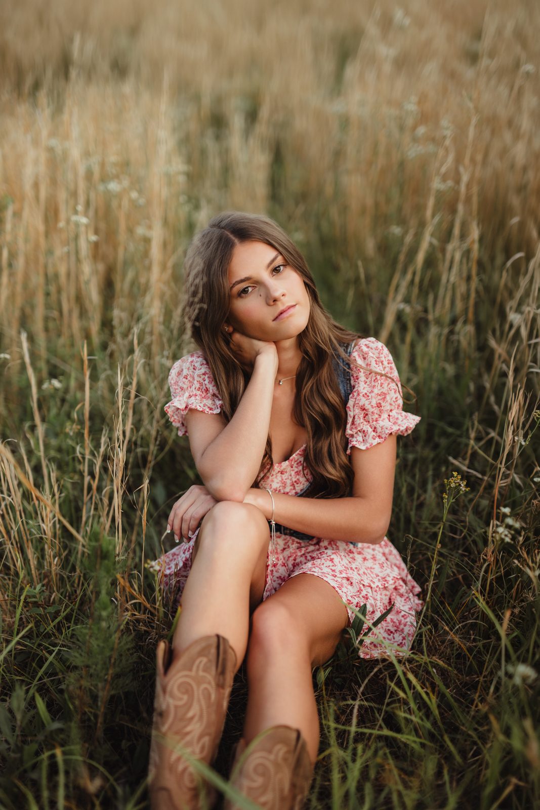 Senior girl in pink floral dress sitting in a field of grass and wearing brown cowboy boots. 