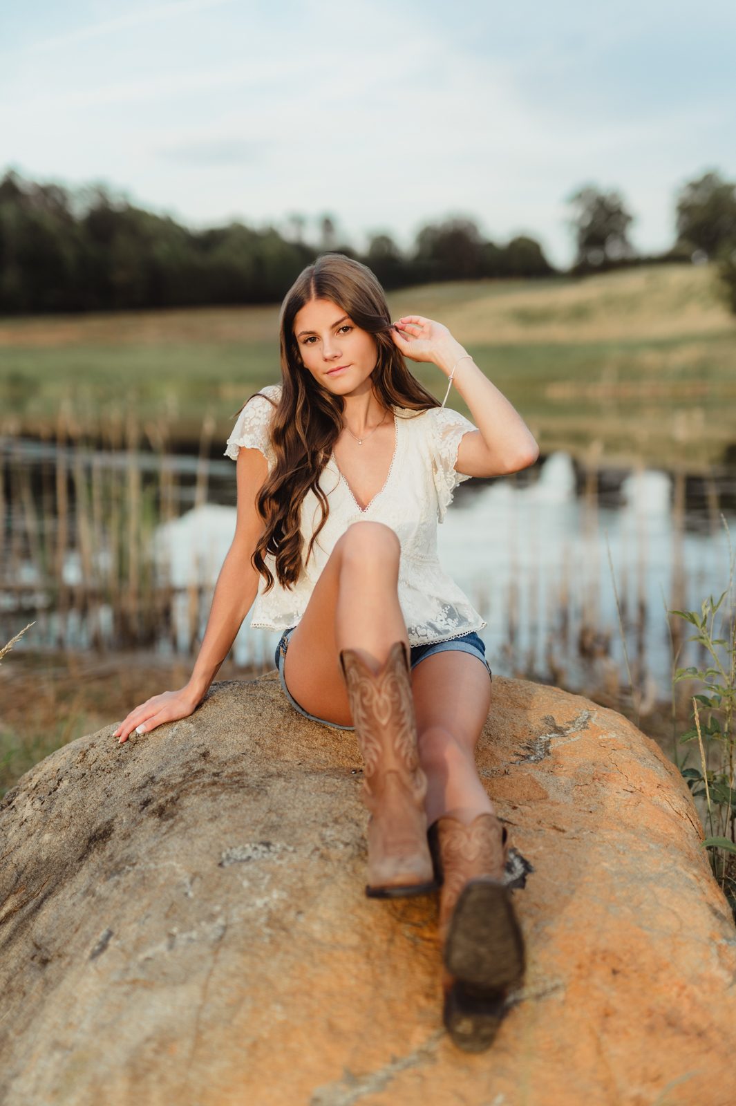 Greensboro senior photographer captures senior girl sitting on a rock by the lake at golden hour, wearing cowboy boots and a lace top