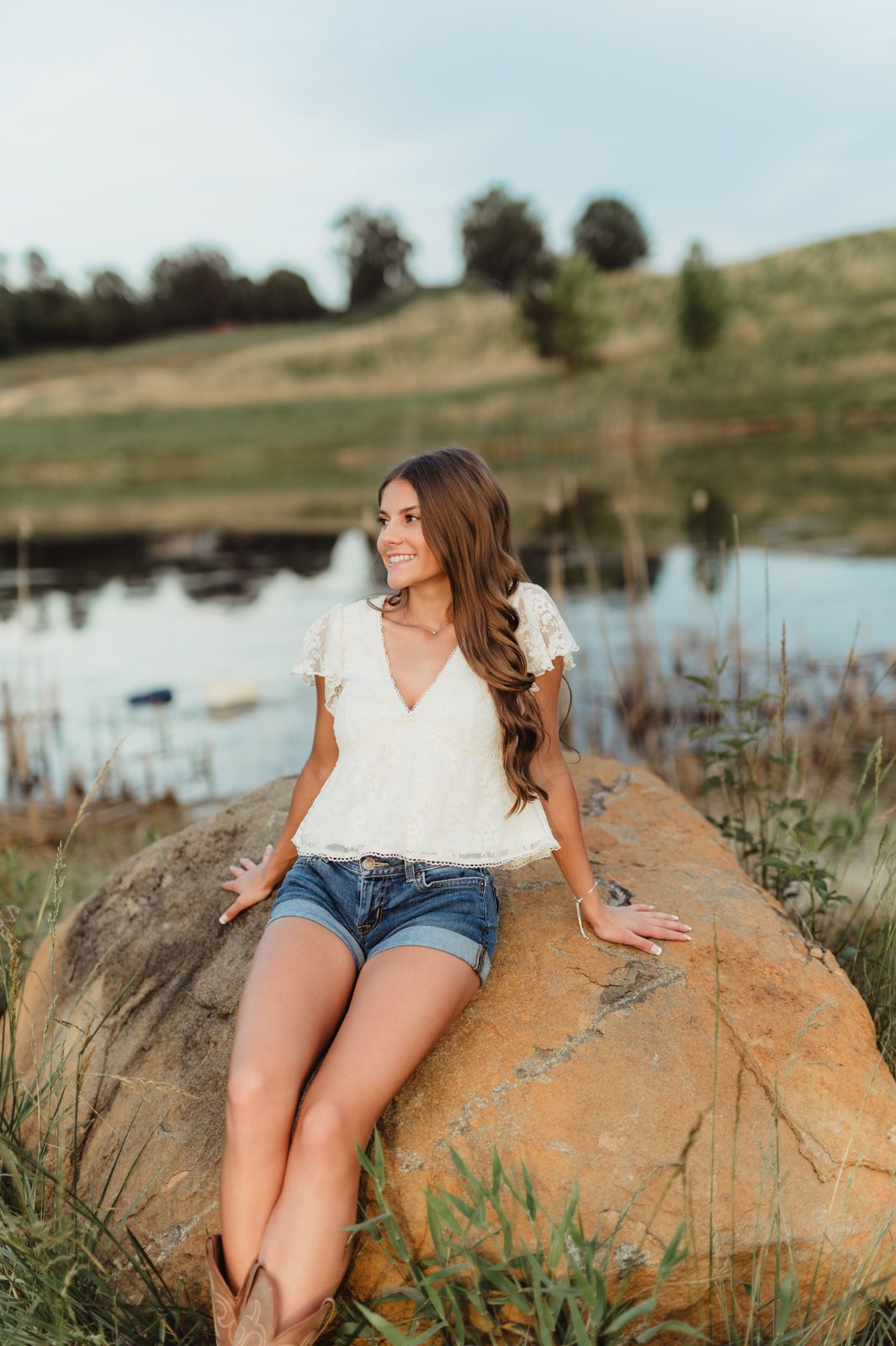 Greensboro senior photo session with a senior girl dressed in white lace top and denim shorts leaning on a lakeside rock at sunset